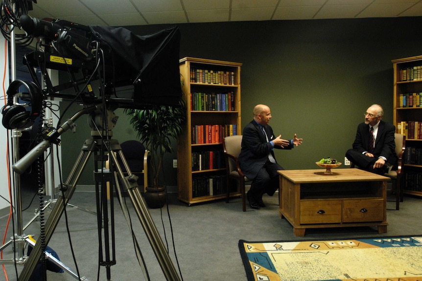 A media training studio with camera equipment, two men at a table, and bookshelves