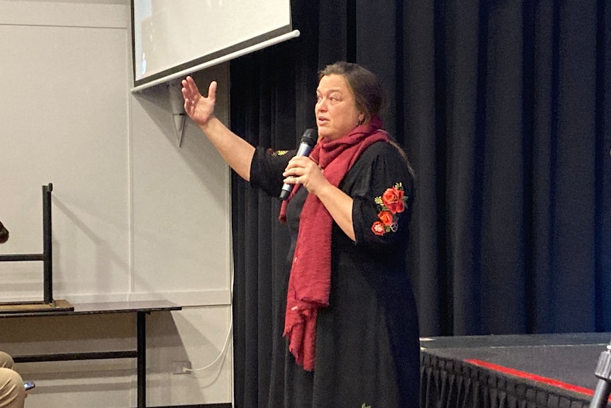 Woman wearing a red scarf gestures as she speaks with a microphone.