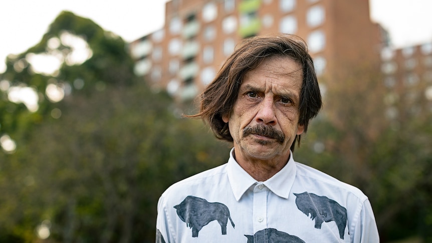 A middle-aged man with a moustache looks directly into the camera, as a tower looms in the background behind him