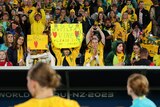 Matildas players watch on at pitchside as Australian fans in the stands cheer and hold up signs after a big match.
