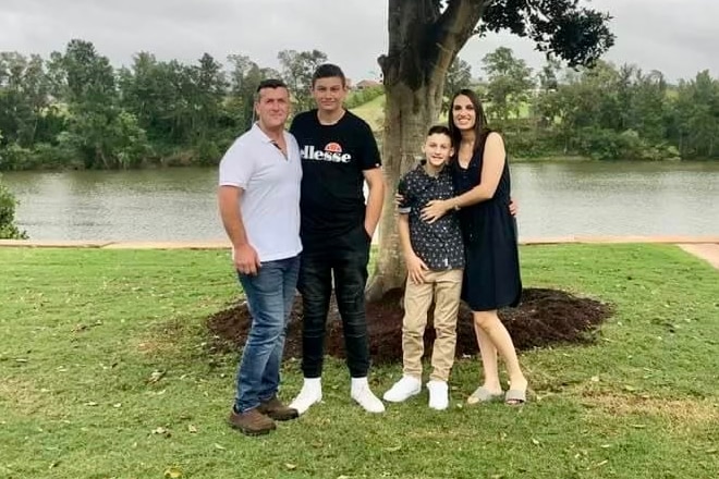 A mother, father and their two sons smiling for a family photo on the bank of a river.