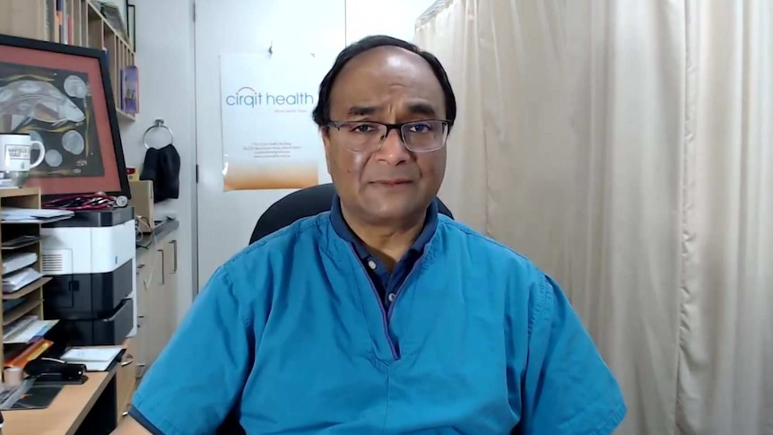 Dr Mukesh Haikerwal sitting at a desk at his clinic, wearing light blue medical top.