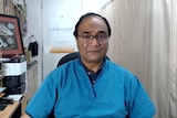 Dr Mukesh Haikerwal sitting at a desk at his clinic, wearing light blue medical top.