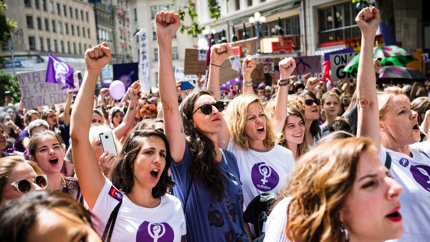 A group of women wearing shirts with purple logos gathered in the streets, their firsts in the air.