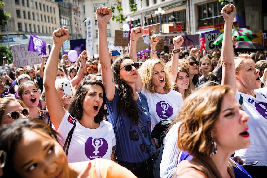A group of women wearing shirts with purple logos gathered in the streets, their firsts in the air.