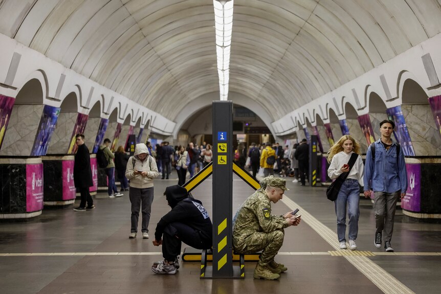 A man in military uniform sits down back to back with a man in a hoodie inside a subway shelter