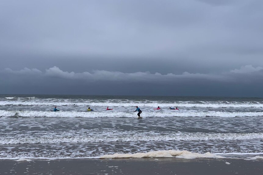 About seven surfers out on the water with grey clouds closing in