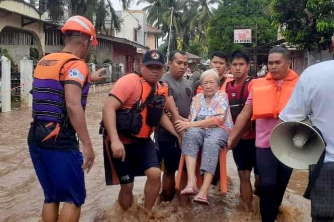 A white-haired lady is carried on a chair by a group of younger men on a flooded street