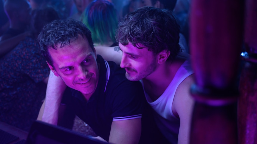A film still of Andrew Scott and Paul Mescal in a busy nightclub. Mescal has his arm around Scott who is leaning over a bar.