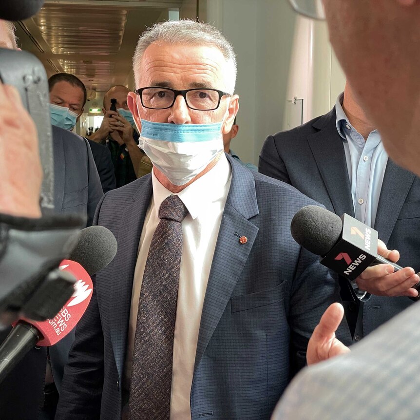 A grey-haired man wearing a mask and glasses speaks to a crowd of journalists and cameras.