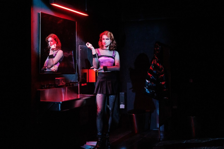 White trans woman with red hair wears pink corset, black skirt and tights and holds vape in neon-lit bathroom.