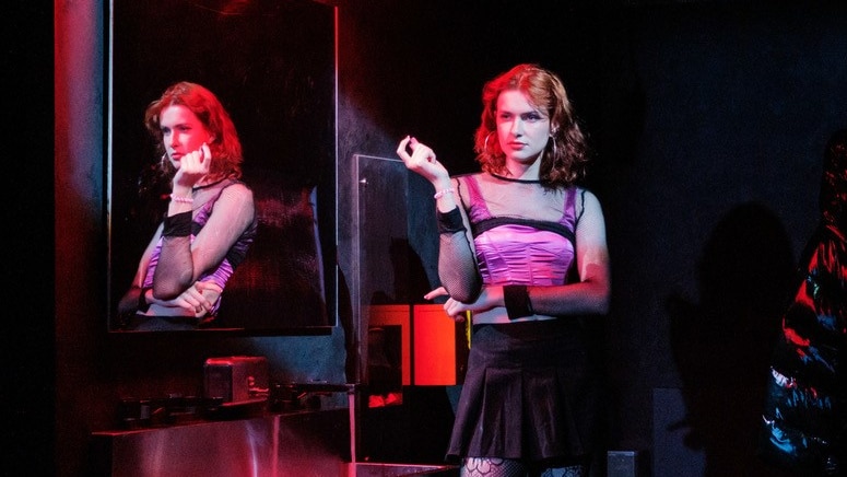 White trans woman with red hair wears pink corset, black skirt and tights and holds vape in neon-lit bathroom.