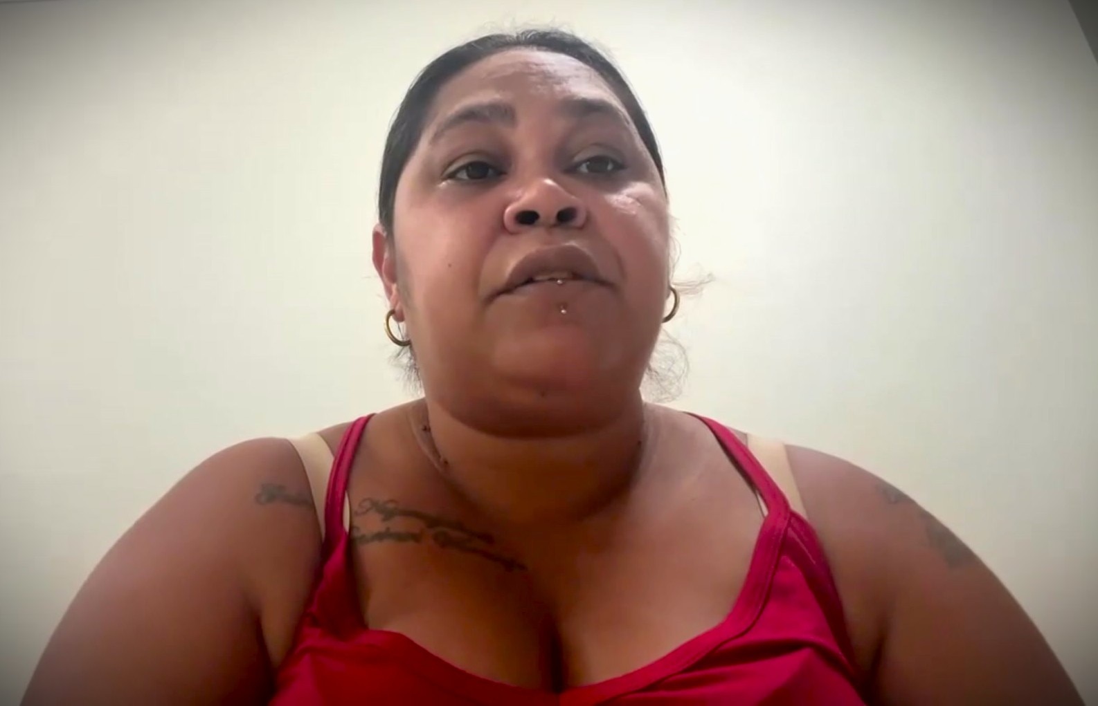 An Indigenous woman, tanned, black hair, looking down at screen, red singlet on, neutral expression.