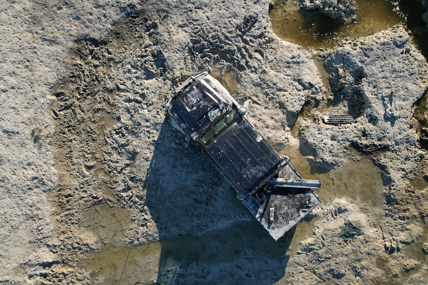 An aerial photo of a ute bogged in thick mud with puddles of water and recovery gear nearby.