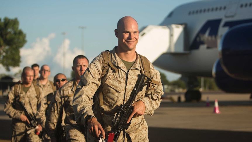 A grinning Marine walks down the airport tarmac, with a gun in his hand
