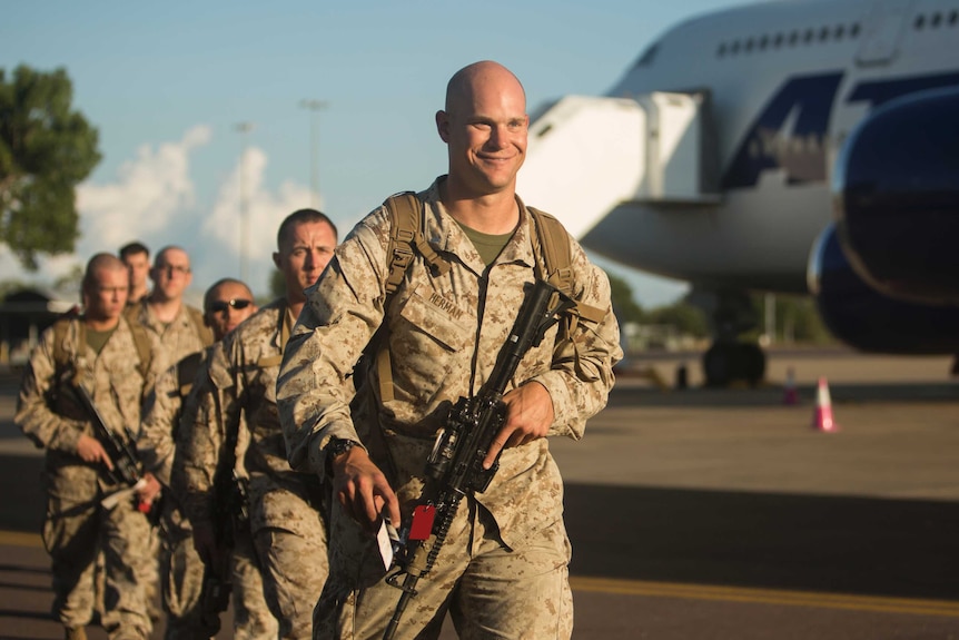 A grinning Marine walks down the airport tarmac, with a gun in his hand