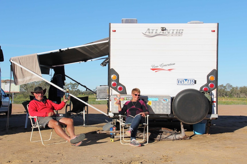 A man and a woman sitting next to a caravan, drinking a wine and beer.