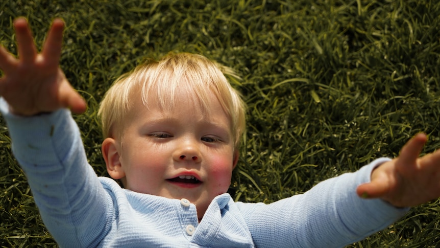 Blonde-haired male toddler smiling with greenery in the background.