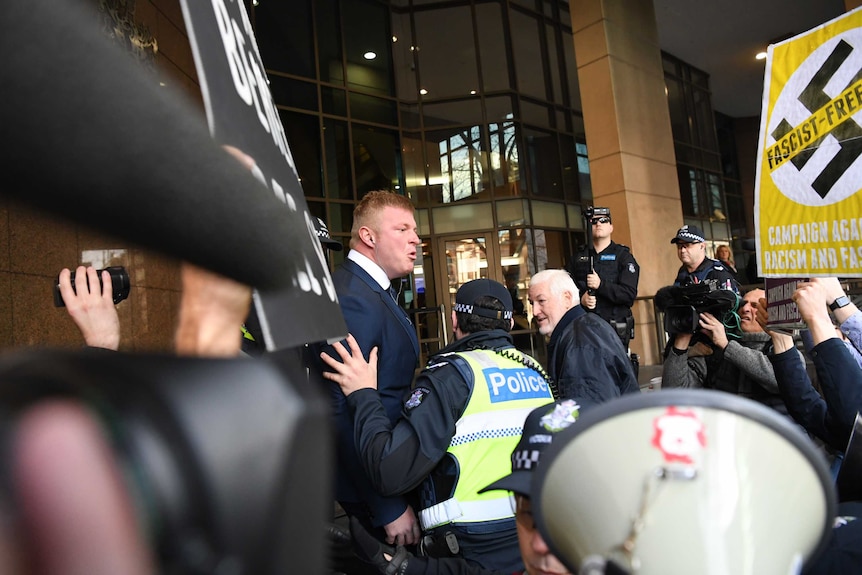 United Patriots Front leader, Blair Cottrell is pushed forward by a policeman with a television camera in the foreground.