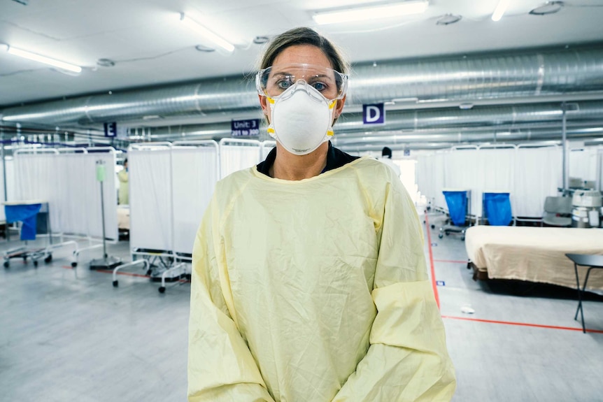A woman in a face mask, goggles, and yellow scrub gown stands in a carpark with hospital beds behind her