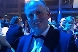 A man in a tuxedo smiles as he holds a trophy while seated at a table at a large gala event.