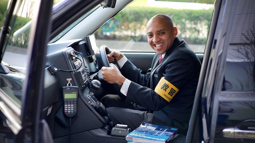 A man beams while sitting in the driver's seat of a Japanese taxi