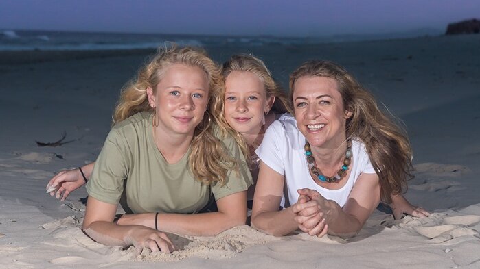 Two young girls and their mother lay on the sand together, smiling. The ocean can be seen behind them.