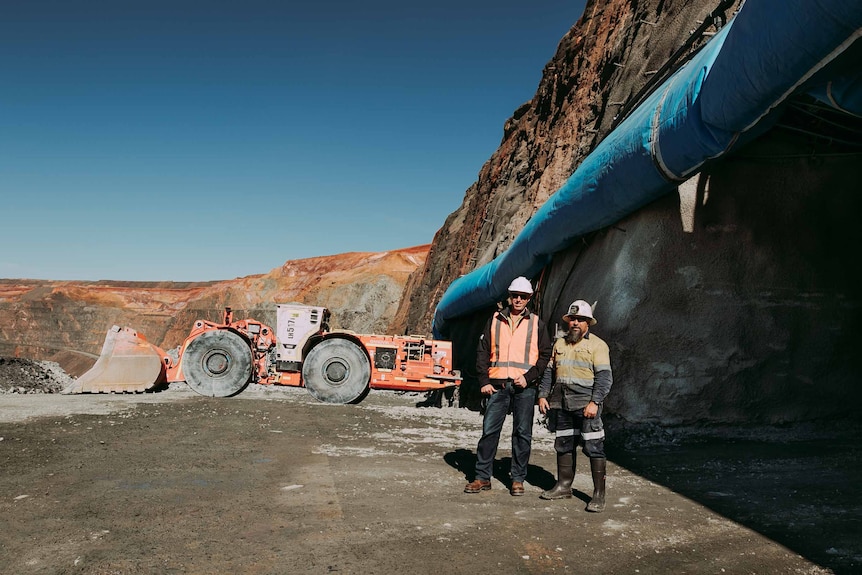 Deux hommes portant des vêtements de travail haute visibilité debout près de machines stationnées dans une mine d'or.  