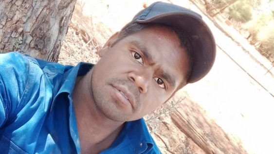 A man wearing a cap and a work shirt takes a selfie on an angle out in the bush.