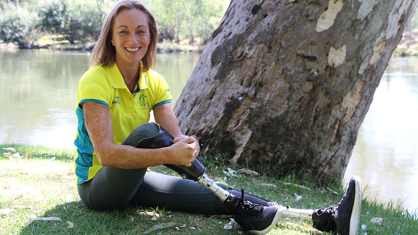 Para-athlete Eliza Ault-Connell sits on grass in front of a tree with river in background.