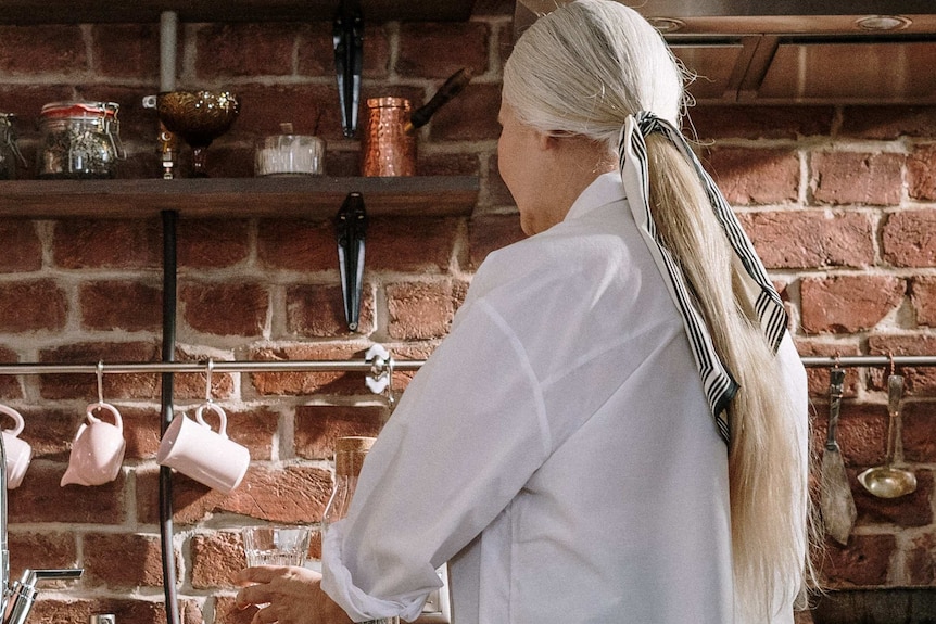 A woman with long grey hair faces away from the camera as she stands at a kitchen bench.