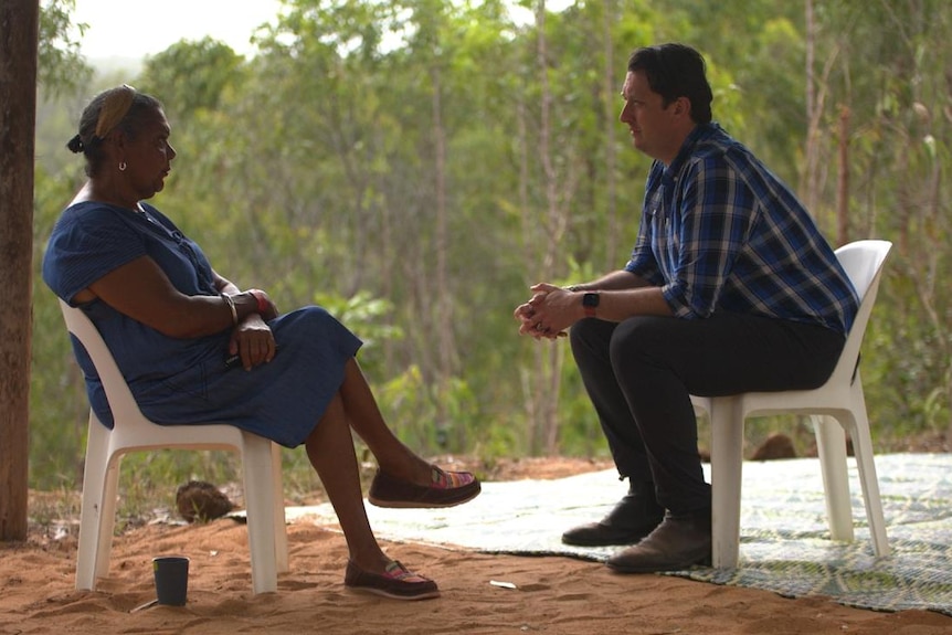 A woman and man sit opposite each other, surrounded by trees.