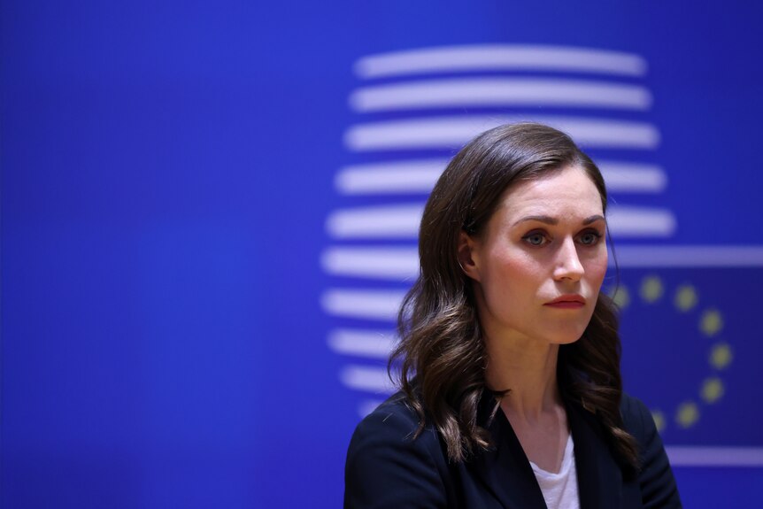 sanna marin looks straight ahead with a serious expression in front of a blue eu banner in the background