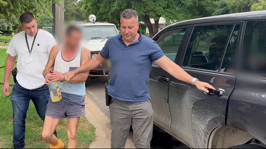 A man in a singlet is escorted into a car by two men.