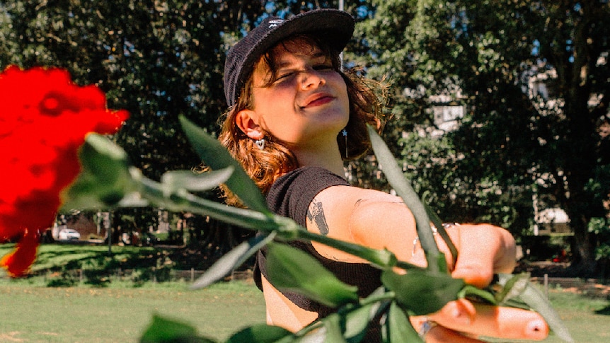 Image of MAY-A, wearing a black cap and top, holding a red flower