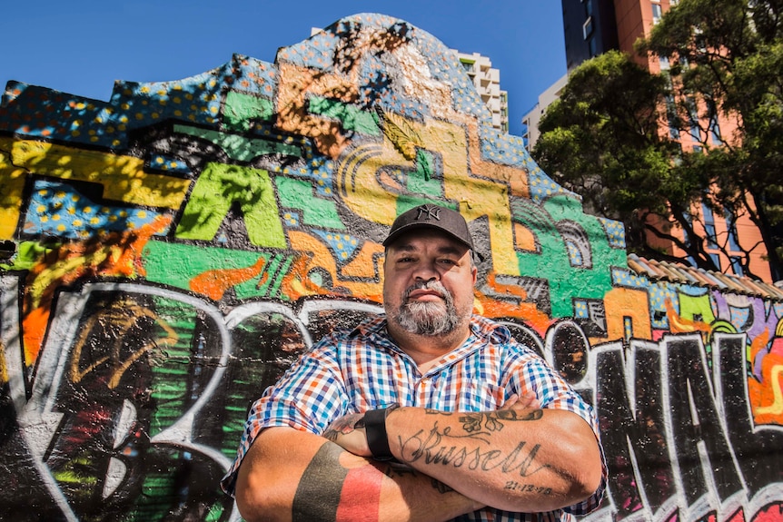 Colour photograph of director Adrian Russell Wills posing in front of a graffitied wall on a sunny day.