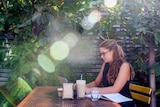 Annie Belcher sits under trees in a cafe courtyard working on her computer, with a smoothie on the table next to her.