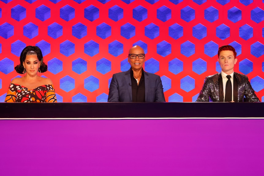 The three people on the panel of judges on the reality TV show RuPaul's Drag Race Down Under sit behind a long table.