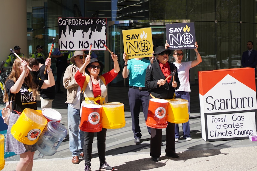 A wide shot of a group of protesters holding signs and drums outside the Woodside building in Perth.