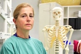 A woman with blonde hair in a ponytail stands beside a skeleton in an office 