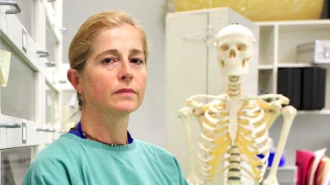 A woman with blonde hair in a ponytail stands beside a skeleton in an office 