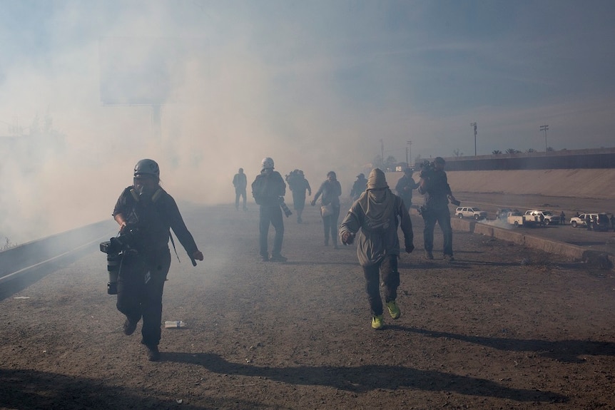 Nine people with covered faces stand on a dirt road as tear gas spreads through the air around them.