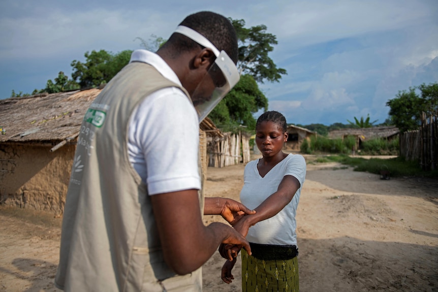 a man with a face shield on checks the arms of a woman standing on the dirt road of a village