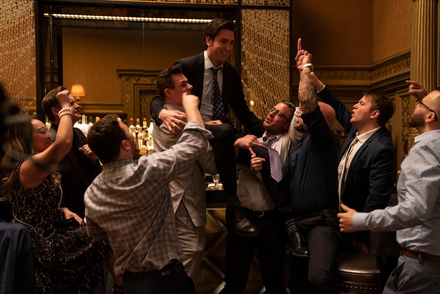 Still from the TV show Succession: a group of mostly men in shirts and suit jackets lifting a young man in a suit up on a chair