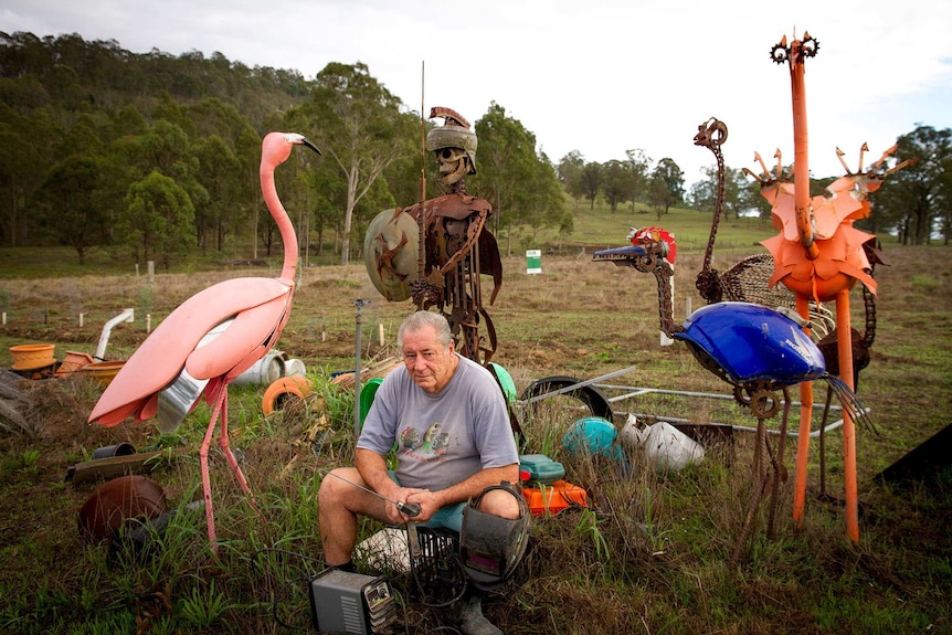 Man sitting in a paddock holding welding equipment surrounded by sculpture made using found objects.