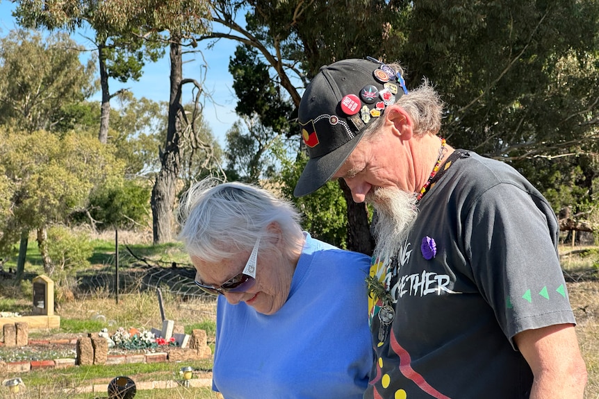 A woman with white hair and a man wearing a cap with Aboriginal badges on it stand side by side with bent heads. 