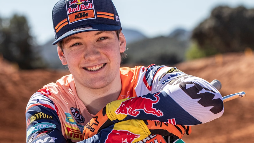 A young man wearing BMX bike riding gear smiles beside his orange and black motorcross bike 