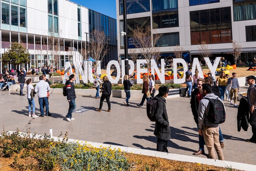 A crowd of young people in an open paved area outdoors near a large sign reading "ANU OPEN DAY".