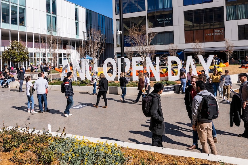 A crowd of young people in an open paved area outdoors near a large sign reading "ANU OPEN DAY".