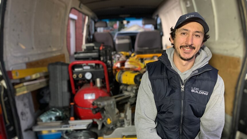 Carpenter Luke Kyriakides sits in the back of a white van wearing a puffer vest and cap, with tools behind him.
